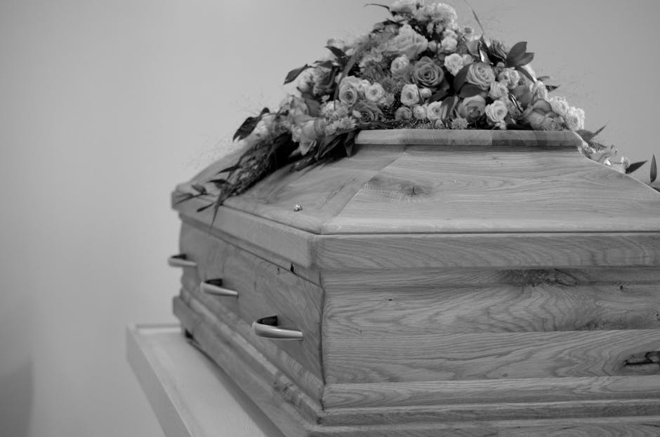 A wooden funeral casket with a polished, natural wood grain finish rests on a white surface. On top of the casket, there is a large, elaborate floral arrangement featuring a variety of roses, lilies,