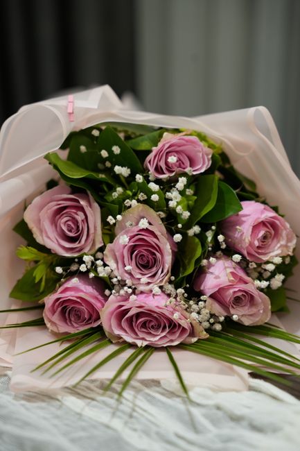 A bouquet of fresh pink roses with delicate, tightly curled petals, accented by small white baby's breath flowers and lush green leaves, arranged in a circular cluster. The bouquet is wrapped in white