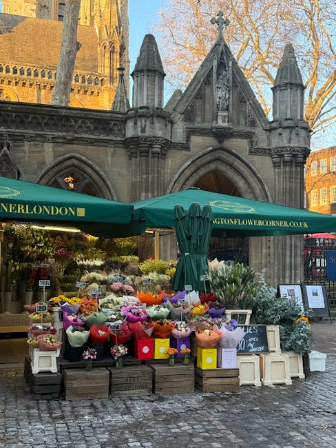 An outdoor flower stall set against the historic stone facade of a church, with pointed arches and detailed gothic architecture, is sheltered by a large green canopy bearing the URL 'langtonflowercorn