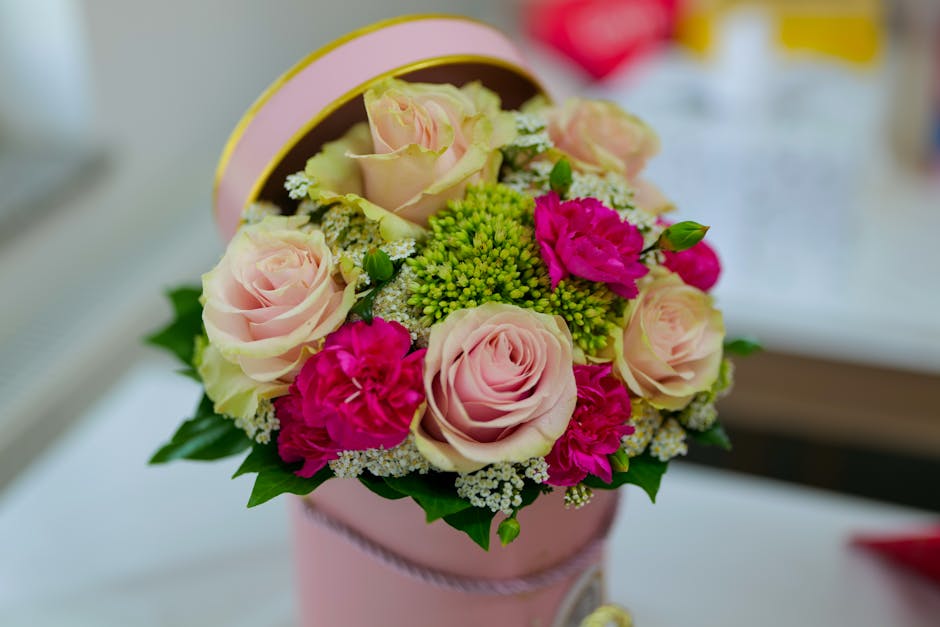 A floral arrangement featuring pale pink roses, vibrant magenta carnations, and small clusters of white filler flowers, all arranged in a round, pink gift box with a decorative ribbon. The bouquet dis