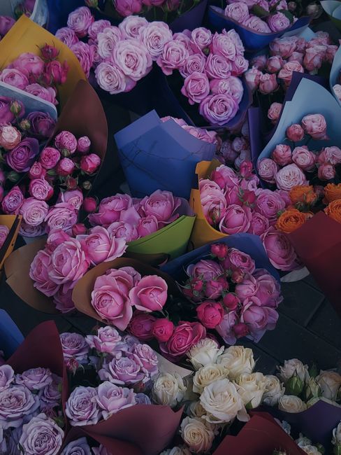 A colorful collection of fresh floral bouquets featuring predominantly pink roses and peonies, arranged in vibrant, pastel-colored wrapping papers including blue, yellow, green, and pink. The bouquets