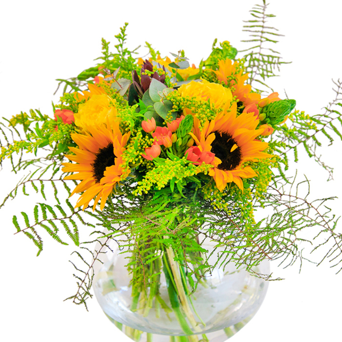 The first impression of this floral arrangement-a true Coulsdon delight-is of unbridled joy and light. Sunflowers stand tall and open, their arching yellow petals and rich brown eyes reminiscent of meadows basking in summer warmth along Marlpit Lane. Tucked among them, butter-gold roses unfold in tightly nested spirals, embodying a sense of luxury and timeless charm. Glossy red Hypericum berries provide vibrant punctuation throughout, evoking the ripening berries seen along local woodlands. Wispy fern fronds and delicately trailing greens cascade around the bouquet, lending movement and a natural, slightly wild essence. Sprigs of goldenrod, studded with tiny yellow blooms, and slender eucalyptus leaves with a hint of blue, appear between the larger flowers, adding soft texture and complexity. The spherical, see-through vase beautifully reveals the bundle of stems, their green hues magnified by the water inside-a testament to Coulsdon Florist's eye for fresh details. Subtle fragrances rise from the bouquet, mingling rose, greenery, and a whisper of sunlit meadow. Crafted to be a joyful gift or an unforgettable centerpiece, this arrangement brings the warmth and beauty of the outdoors right into a Coulsdon living room-perfect for any occasion that calls for a burst of happiness and colour.