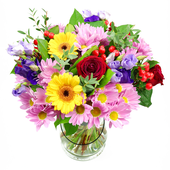 A full, radiant bouquet bursts from a clear glass vase, its colours and textures heightened by a clean white backdrop that draws the eye straight into the flowers. At its heart, sumptuous deep red roses form a rich, velvety core, their layered petals curving inward with classic elegance, suggesting heartfelt affection or a meaningful anniversary surprise somewhere in residential Coulsdon. Framing these roses are two striking yellow gerbera daisies, their dark brown-orange centres and bold petals reminiscent of miniature suns, instantly brightening the composition with a sense of friendliness and joy. Encircling the central blooms, plentiful soft pink daisies or chrysanthemums gather in loose clusters, each flower formed of many slender petals that catch the light and create a soft, airy volume. Their warm yellow centres add a gentle glow, as if echoing the warmth of a cosy local café near Chipstead Valley Road. Threaded between the pinks and yellows, purple and lavender lisianthus emerge on graceful stems, their bell-shaped flowers and cool tones giving the design depth, sophistication, and a hint of calm. Small, rounded red hypericum berries are tucked throughout the bouquet, adding an unexpected texture and a contemporary twist to the otherwise classic selection of blooms. Lush greenery interlaces everything-broad, dark leaves that ground the design, along with finer, pale-green shoots that bring lightness and movement-creating the impression of a bouquet freshly gathered from a well-loved garden. The clear vase reveals straight, evenly cut stems resting in clear water, underlining the florist's care and precision. This arrangement feels ideal for birthdays, thank-you gifts, or simply brightening a Coulsdon living room with a burst of colour and gentle, uplifting fragrance.